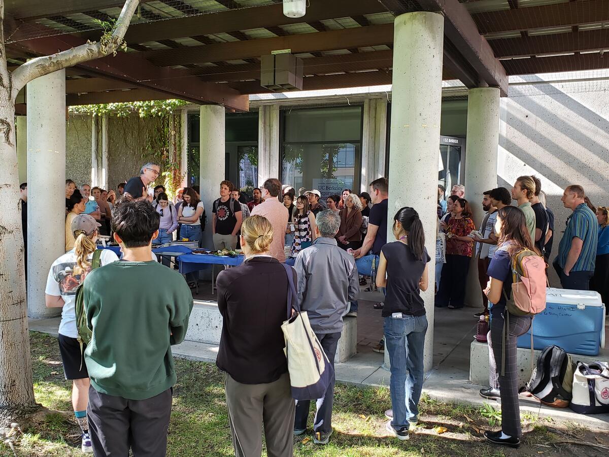 Corey Goodman speaking to a crowd of dozens of people standing around him, with a hand on his chest. He is facing Dan Feldman. They are outside, directly in front of Barker Hall, and there is a table of food in the middle.