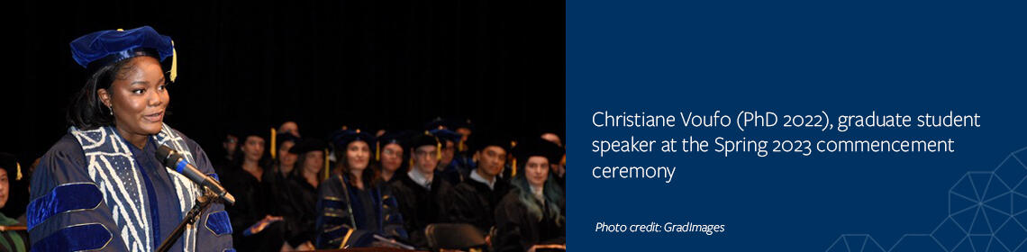 Christiane at left of image, standing at podium on stage with seated graduates in the background.